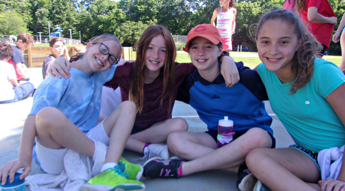 Four female campers of middle school age sitting on the sidewalk together with their arms around one another and smiling