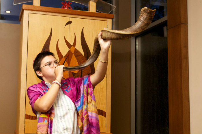 Individual wearing a pink prayer shawl and blowing a shofar in a synagogue sanctuary