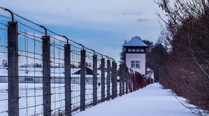 Dachau concentration camp is visible in the distance behind a long barbed wire fence 
