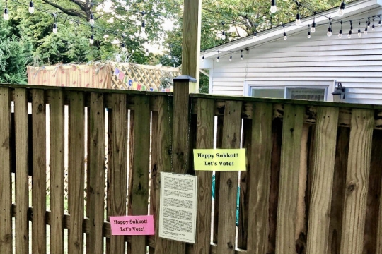 Fence with a printed letter on it and two signs reading ITS SUKKOT LETS VOTE with a sukkah and house visible in the background