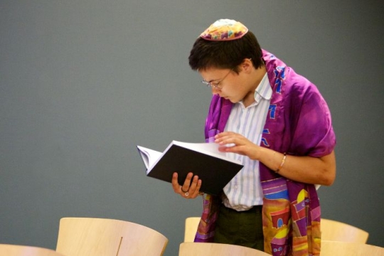 Person wearing a prayer shawl and kippah reading a prayer book in pews alone